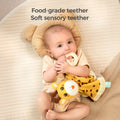 A baby lying on a bed with a bear-shaped pillow, holding and chewing on a food-grade teether attached to a giraffe baby security blanket. The text above says, 