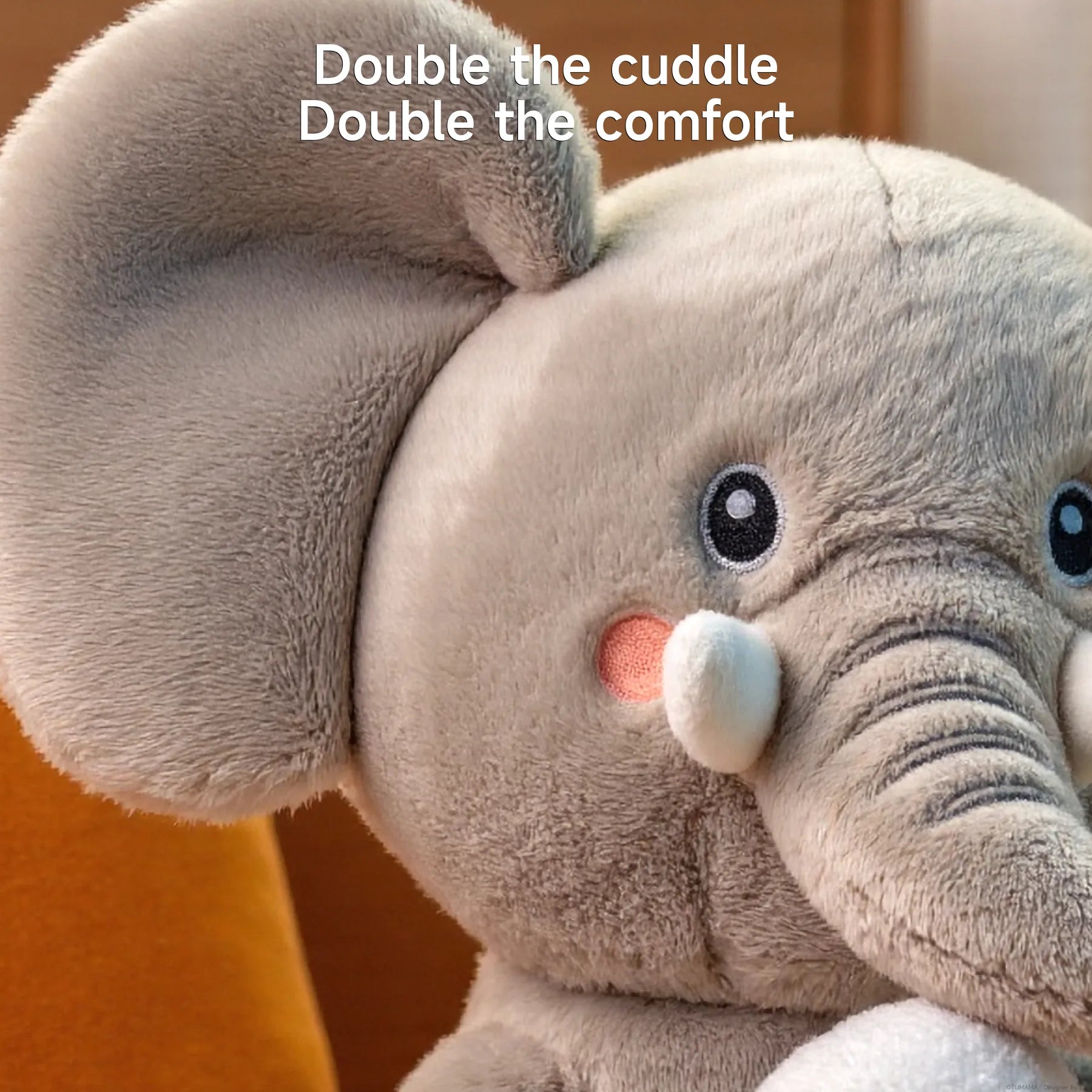 A close-up view of the plush elephant toy shows its ultra-soft, fuzzy texture, friendly embroidered eyes, blushing cheeks, and gentle trunk. Text at the top reads, “Double the cuddle, Double the comfort,” emphasizing the toy’s softness and huggability.