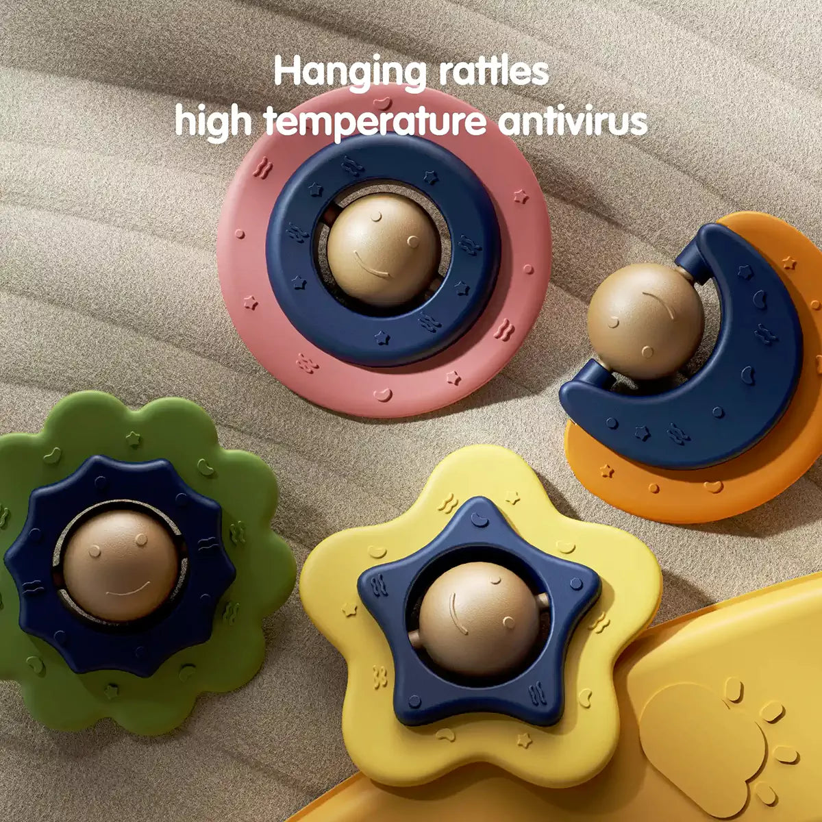 Close-up view of four colorful baby rattles designed for hanging on a play gym. The rattles are shaped like a pink circle, green flower, yellow star, and orange crescent moon, each with a smiling face in the center. The surface is textured with small embossed patterns. 