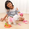 Young girl sitting on the floor playing with a white plush dog toy in a pink lace dress, holding a toy hairdryer as part of a pretend pet grooming set, with accessories like a brush, mirror, stethoscope, and dog bowl nearby.