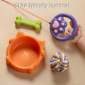Close-up of child-friendly toy accessories including a cat-shaped orange food bowl, green bone, multicolored rope ball, and a purple paw-shaped remote held by a child's hand, designed for interactive pet play.