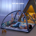 Baby sleeping peacefully on a soft cotton play mat with grey arches, under hanging plush rattles including an elephant, lamb, bear, and dog. Side cloth book panel with colorful animal illustrations, cozy tent setup in the background.