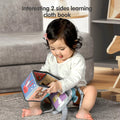 Baby sitting on a carpet, playing with a colorful two-sided cloth book featuring animal illustrations, smiling and engaged in learning activity.